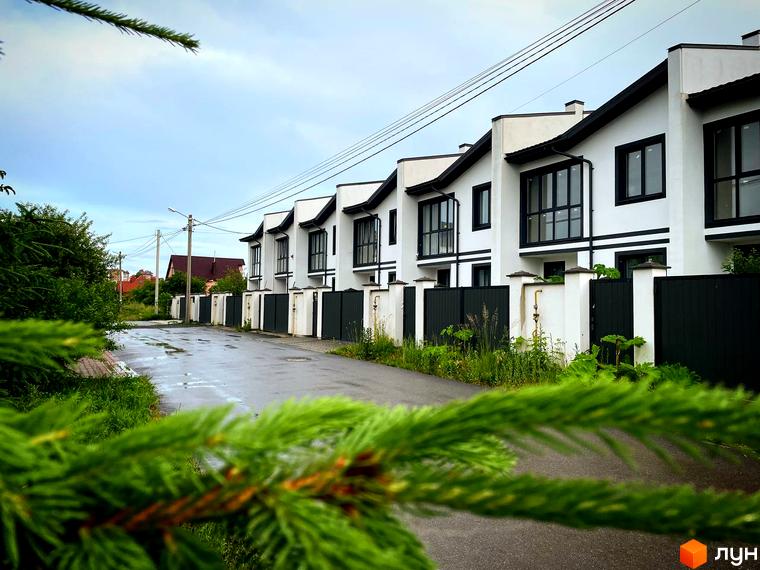 Ряд білих таунхаусів WEST TOWN I з чорними дахами та віконними рамами. Перед будинками мокра дорога, огорожа та зелені насадження.