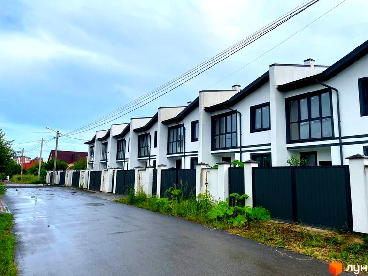 Ряд таунхаусів WEST TOWN I білого кольору з чорними дахами та віконними рамами. Перед будинками розташована дорога, зелені насадження та огорожа.