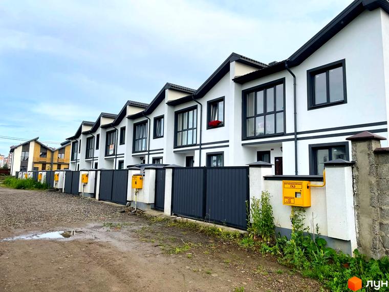 Ряд білих таунхаусів Таунхаус WEST TOWN III з темними дахами та віконними рамами. Перед будинками розташовані огорожі та ґрунтова дорога з рослинністю.