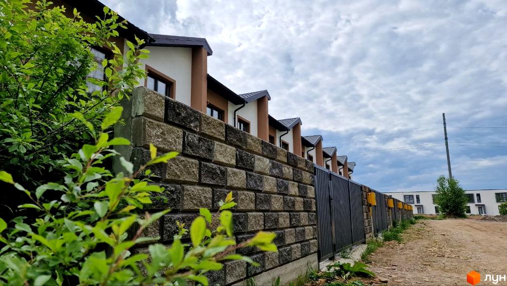 Фото фасаду таунхаусів WEST TOWN IV за парканом з цегли. Навколо будівель зелені насадження та дорога.