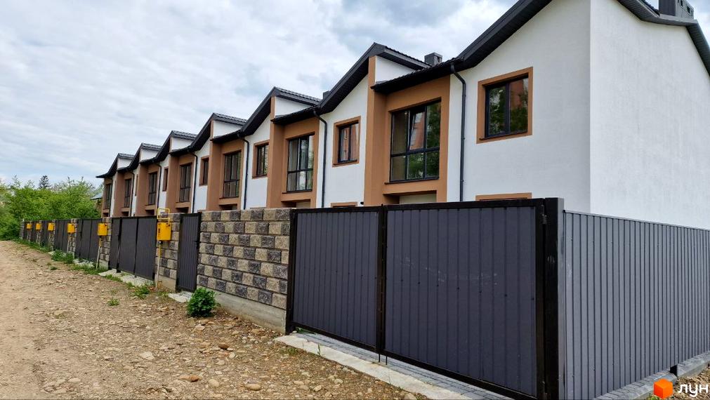 Фасад Таунхаус WEST TOWN IV з білими та коричневими стінами, темними дахами та вікнами. Перед будинками розташований паркан та ґрунтова дорога.