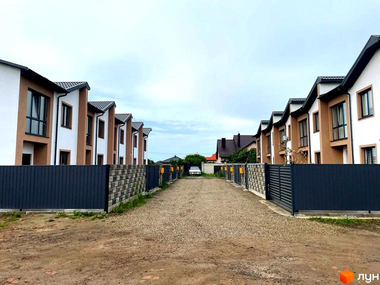 Зображення таунхаусів WEST TOWN V: ряд двоповерхових будинків з білими та коричневими фасадами, розташованих вздовж гравійної дороги з темними огорожами.