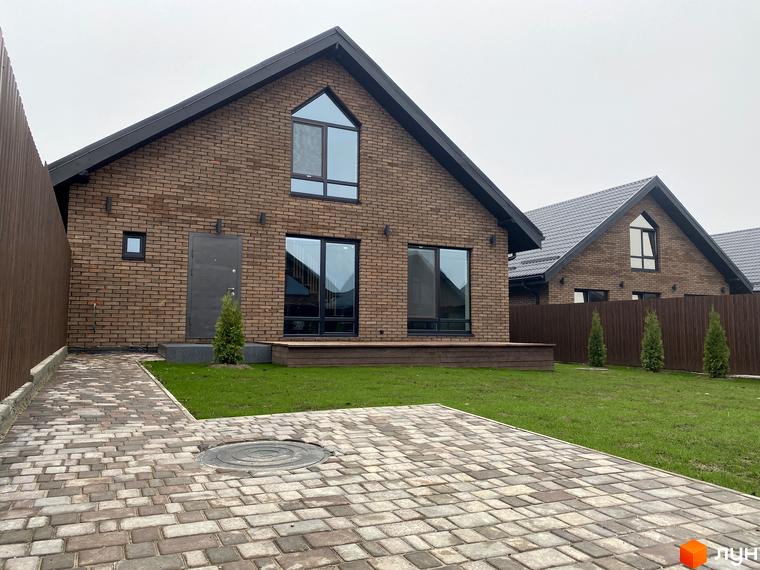 Вигляд котеджу з цегли з великими вікнами в Котеджному містечку Західна Баварія. Навколо будинку зелений газон та вимощена бруківкою доріжка.