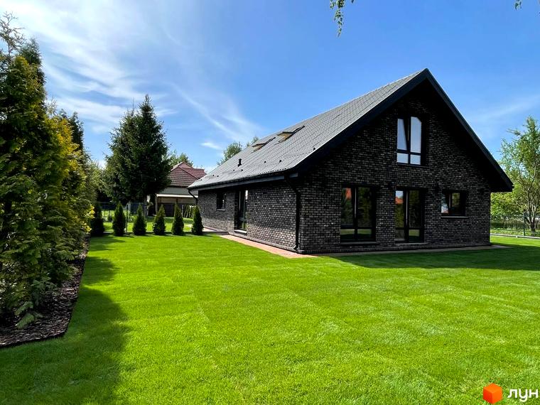 Котеджне містечко Західна Баварія: Фасад будинку з темної цегли та чорним дахом, оточений доглянутим зеленим газоном та вічнозеленими деревами.