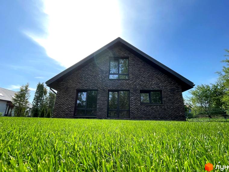 Темний цегляний будинок з великими вікнами та двосхилим дахом в Котеджному містечку Західна Баварія. На передньому плані видно доглянутий зелений газон.