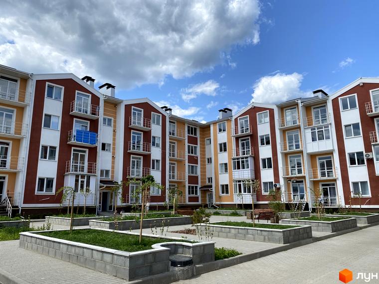 Житловий комплекс Амстердам з фасадом у коричнево-білих тонах, з балконами та панорамними вікнами. На прибудинковій території висаджені дерева та клумби з газоном.
