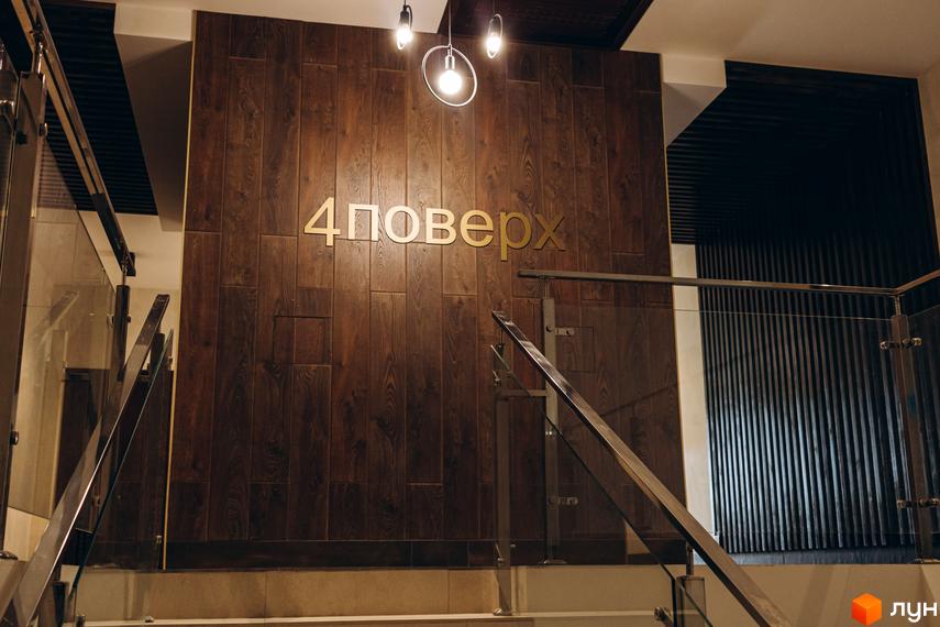 Вхідна група клубного будинку Rezydencja Zalizna Voda з дерев'яною обшивкою стін та золотими літерами назви проєкту. Сучасні перила зі скла та металу ведуть до сходів.