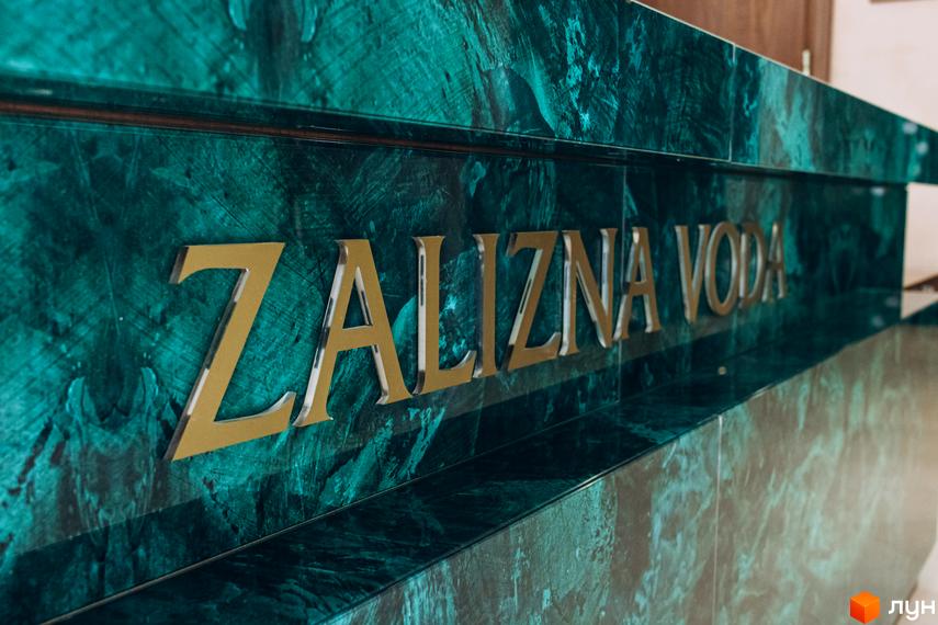 Вивіска клубного будинку Rezydencja „Zalizna Voda” з золотистими літерами на тлі зеленої мармурової поверхні. На задньому плані видно частину інтер'єру.