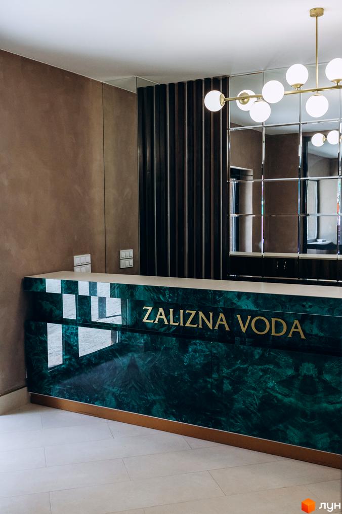 Стильна рецепція клубного будинку Rezydencja „Zalizna Voda“ з мармуровою стійкою та золотим логотипом. Інтер'єр доповнений дзеркалами та сучасною люстрою.