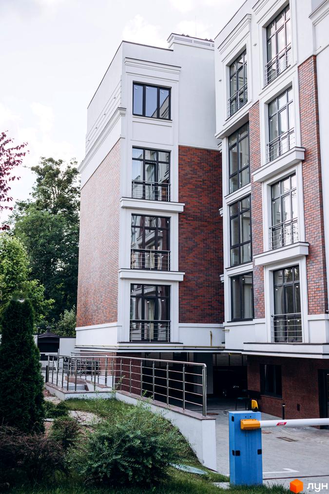 Зовнішній вигляд клубного будинку Rezydencja „Zalizna Voda“ з цегляним фасадом та панорамними вікнами. На прибудинковій території зелені насадження та паркан.