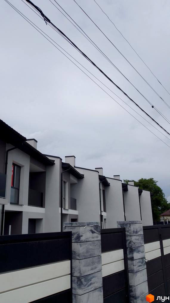 Ряд світло-сірих таунхаусів з темними дахами та балконами. На передньому плані видно паркан та лінії електропередач.