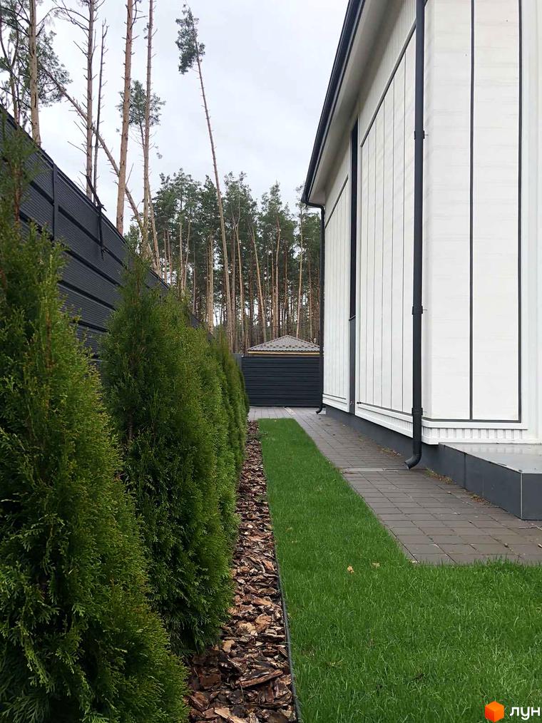 Білий будинок з вертикальними лініями на фасаді та чорними елементами декору, доглянута прибудинкова територія з газоном та туями.