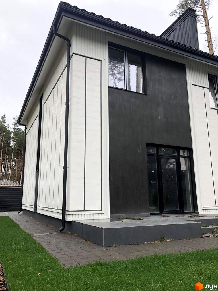 Білий будинок з темними вставками, чорним дахом та водостічною системою. Перед будинком вимощена доріжка з бруківки, газон з травою.