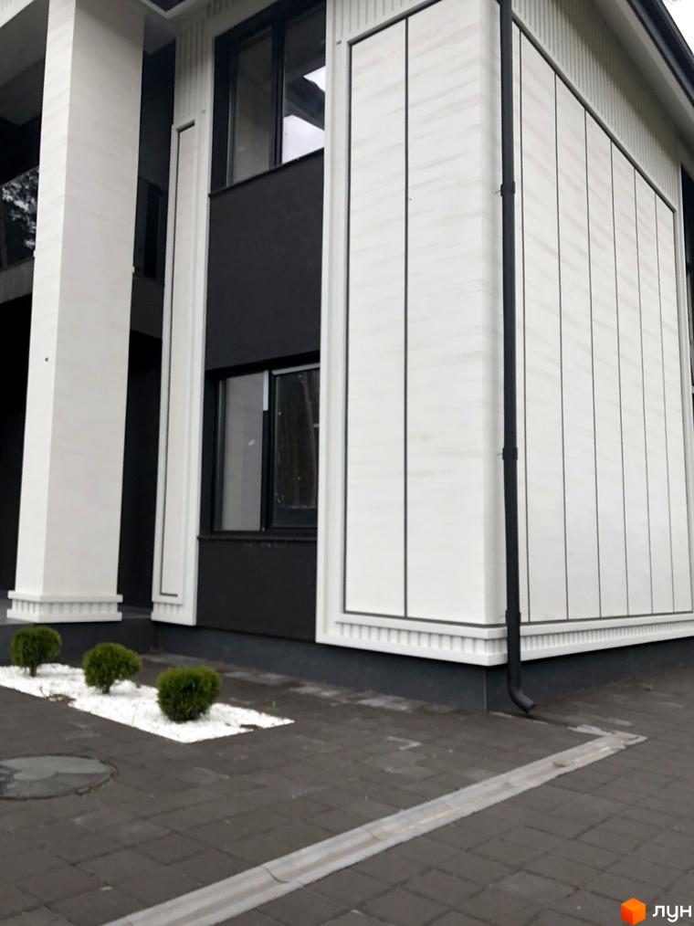 Зовнішній вигляд дуплексів Toscana Home з білими колонами та сірою обшивкою, на тлі доглянутої прибудинкової території з кущами та бруківкою.