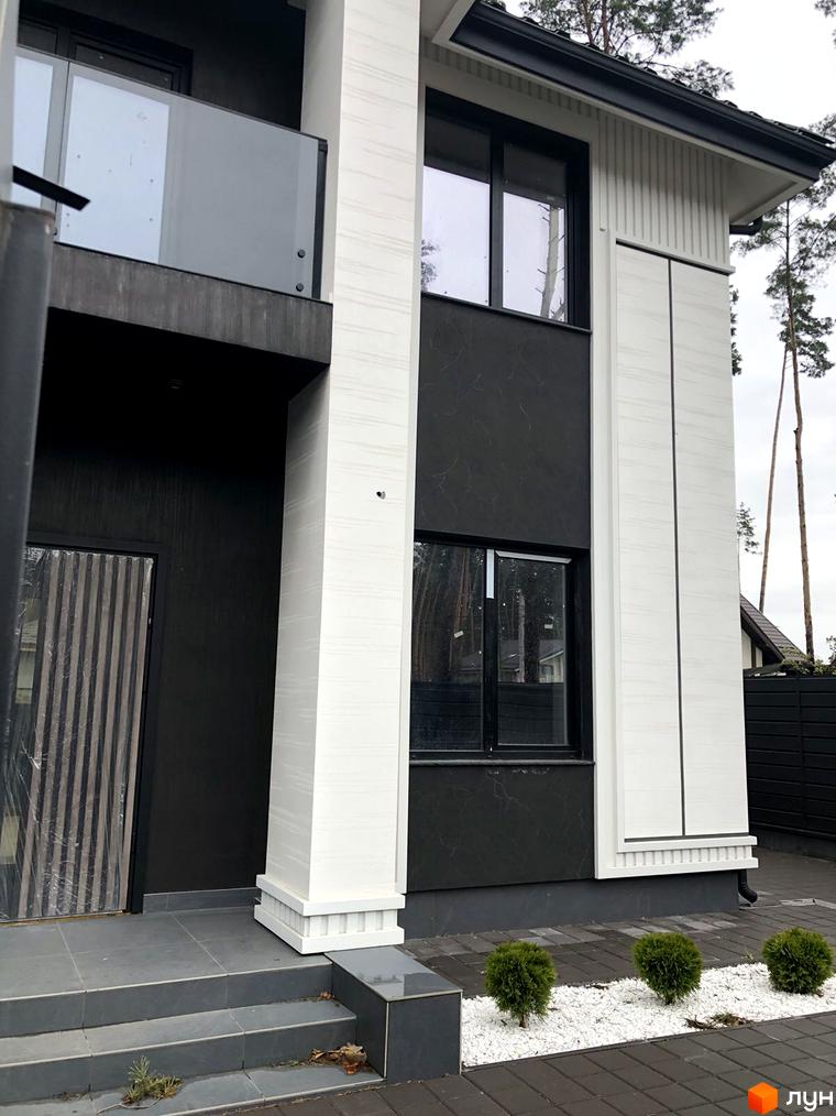 Фасад будинку Toscana Home з білими та темними елементами, скляним балконом та вхідною групою. На прибудинковій території розташовані зелені кущі та доріжка з плитки.