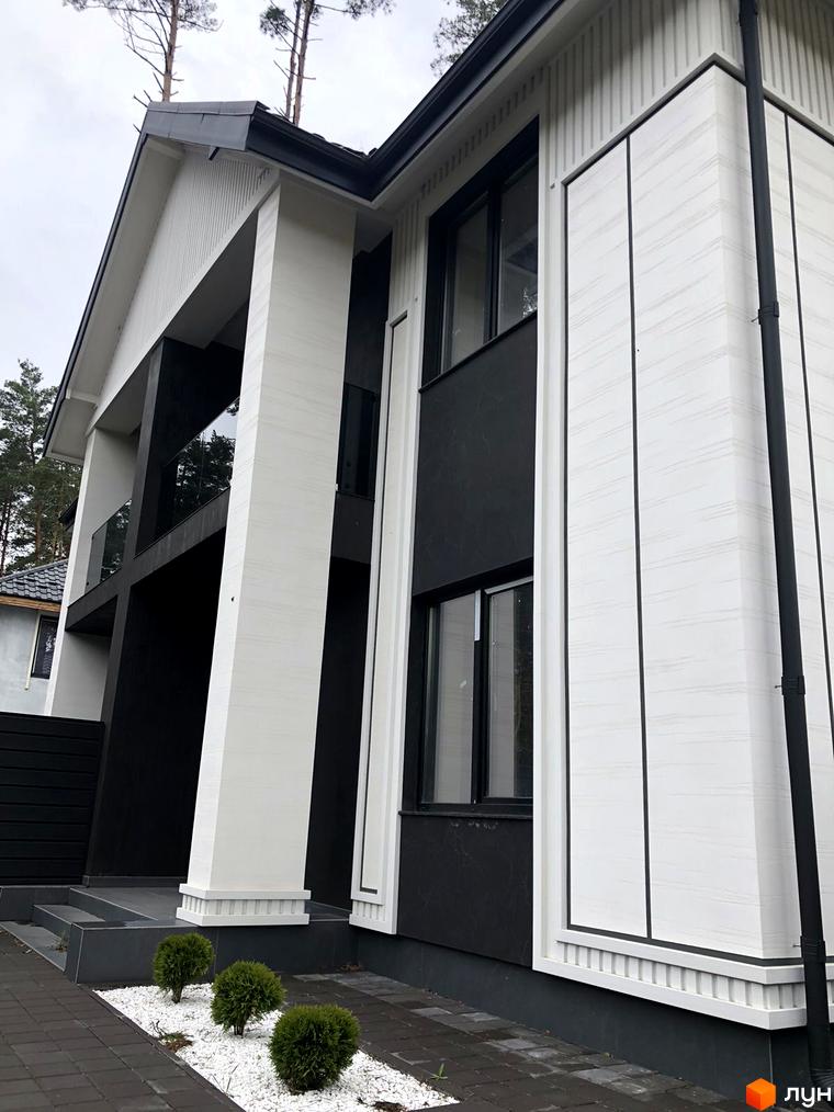 Фасад будинку Toscana Home з білим та чорним оздобленням, великими вікнами та скляним балконом. Навколо будинку доглянута прибудинкова територія з доріжками та зеленими насадженнями.