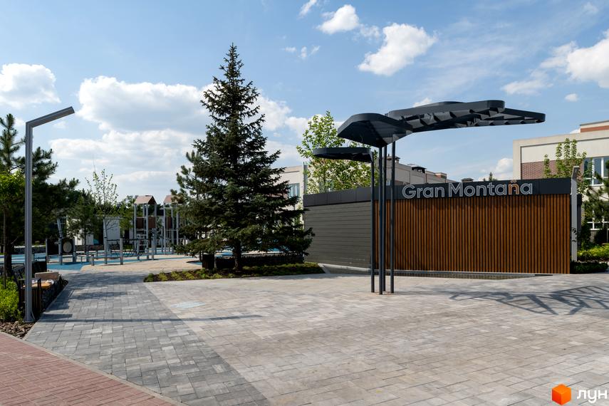 Зображення входу до Котеджного містечка Gran Montana з дерев'яною обшивкою та великим деревом поруч. На території облаштовано пішохідні доріжки.