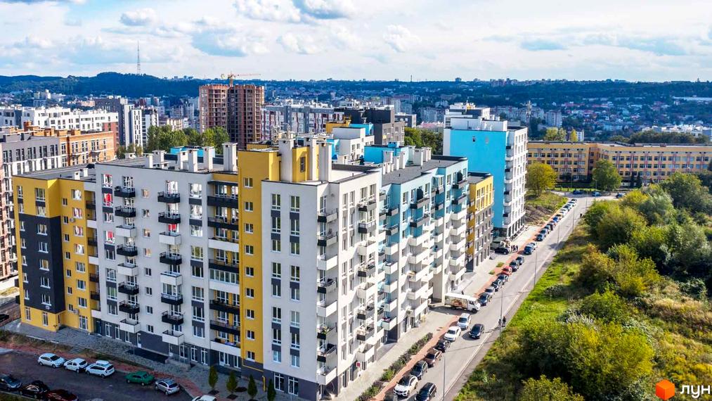 Новобудова ЖК Білий Шоколад з фасадами білого, жовтого та блакитного кольорів, розташована вздовж дороги з припаркованими автомобілями та зеленою зоною.