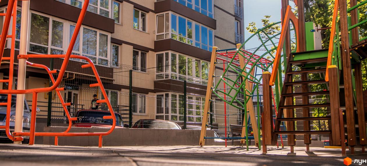 Житловий комплекс Шоколад: Світлий багатоповерховий будинок з коричневими елементами та великими вікнами. На передньому плані дитячий майданчик.