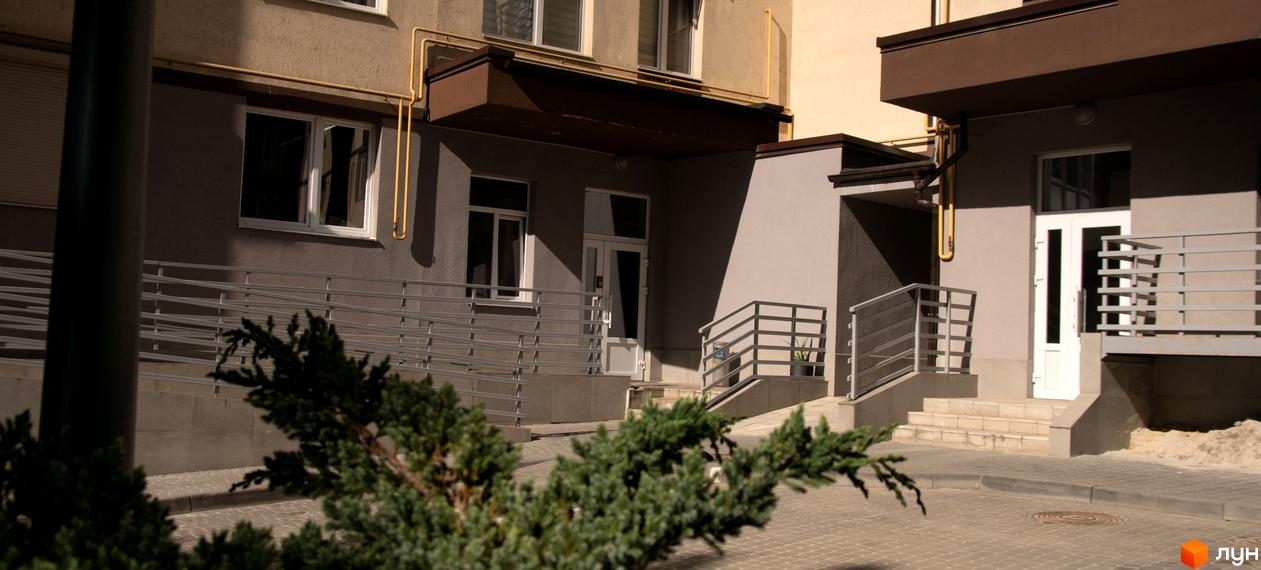 Фасад Житлового комплексу Шоколад з сірими стінами, білими вікнами та коричневими елементами. Прибудинкова територія має пандус, сходи з поручнями та бруківку.