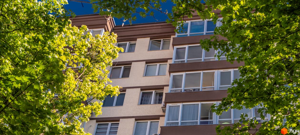 Новобудова Шоколад: багатоповерховий будинок з бежевим фасадом та коричневими елементами, оточений пишними зеленими деревами під синім небом.