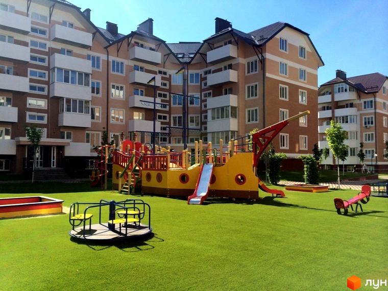 Житловий комплекс Квартал Буча. Багатоповерхові будинки з балконами та дитячий майданчик з пісочницею, каруселлю та гірками на зеленій зоні.