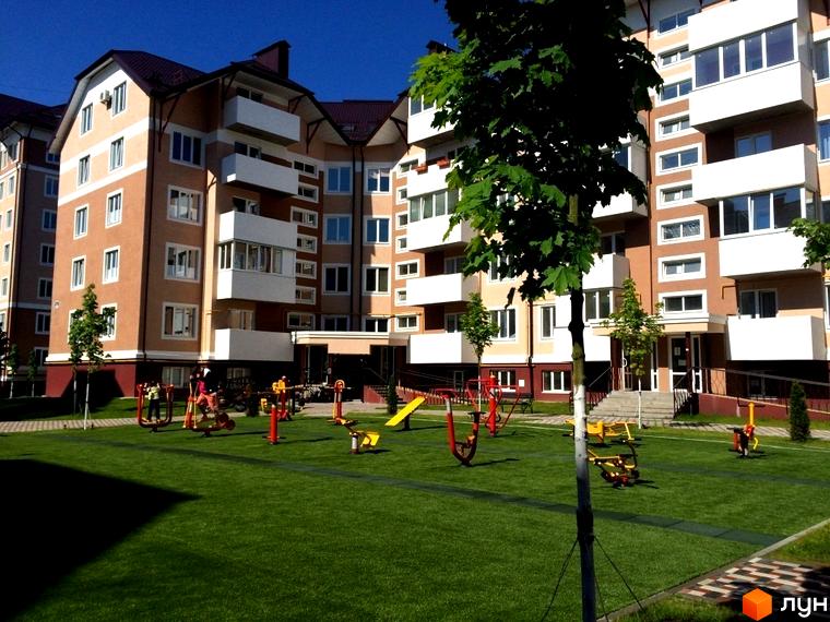 Житловий комплекс Квартал Буча з фасадами світло-бежевого та коричневого кольорів. На прибудинковій території розташований дитячий майданчик та спортивні тренажери на зеленій зоні.