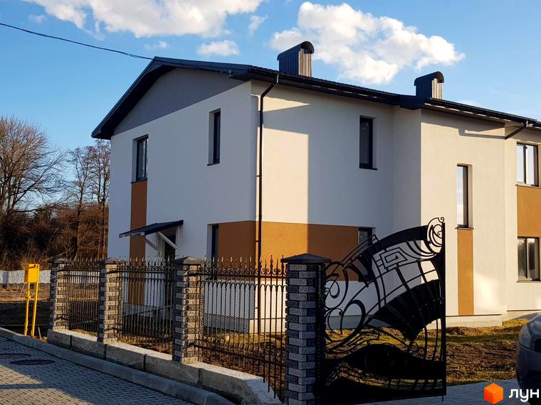 Зображення двоповерхового будинку в Котеджному містечку Горіхова Долина з чорним дахом та металевою огорожею. Навколо будинку зелена трава та дерева.