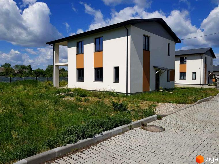 Двоповерховий будинок з білими стінами та коричневими вставками в Котеджному містечку Горіхова Долина. Перед будинком газон з травою та тротуарна плитка.