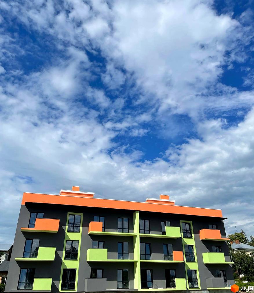Зображення фасаду багатоповерхового будинку ЖК Долина Троянд з помаранчевим дахом та зеленими і помаранчевими вставками на сірому фоні неба з хмарами.