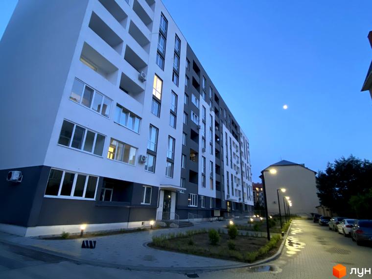 Зовнішній вигляд житлового комплексу Щасливий з білими та сірими фасадами, балконами та великими вікнами. Прибудинкова територія з освітленням та доріжками.