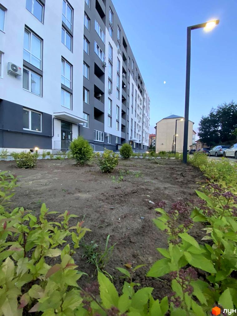 Зовнішній вигляд житлового комплексу Щасливий з біло-сірими фасадами та кондиціонерами, прибудинкова територія з зеленими насадженнями та освітленням.