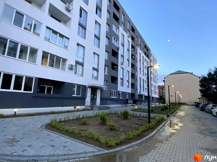 Зовнішній вигляд новобудови ЖК Щасливий з сірими та білими фасадами, вікнами та кондиціонерами. Прибудинкова територія з вимощеною доріжкою, освітленням та зеленими насадженнями.