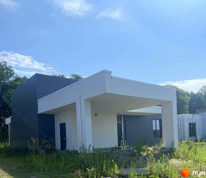 Котеджне містечко Нова Конча-Заспа CRYSTAL. Зовнішній вигляд будинку з білою навісною частиною та темною стіною, на тлі зеленої території та блакитного неба.