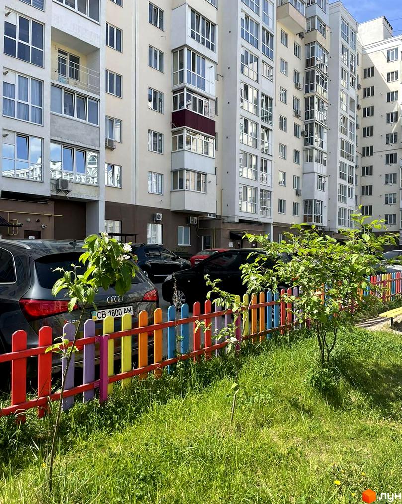 Зовнішній вигляд багатоповерхового житлового комплексу на Масанах. Прибудинкова територія з автомобілями та кольоровою огорожею.
