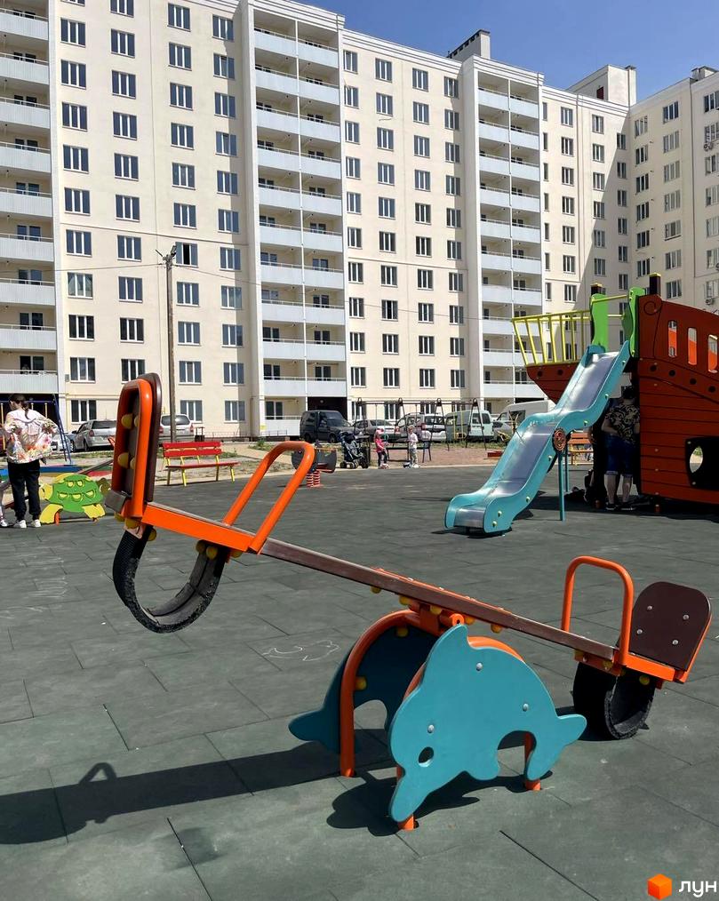 Новобудова ЖК на Масанах з дитячим майданчиком. Багатоповерховий будинок світлого кольору з балконами та дитячими гойдалками, гіркою та іншими елементами для відпочинку.