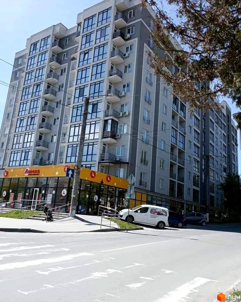 Висока будівля ЖК Фортеця з великою кількістю вікон та балконів. На першому поверсі розташований магазин з жовтою вивіскою, перед будинком пішохідний перехід та дорога.