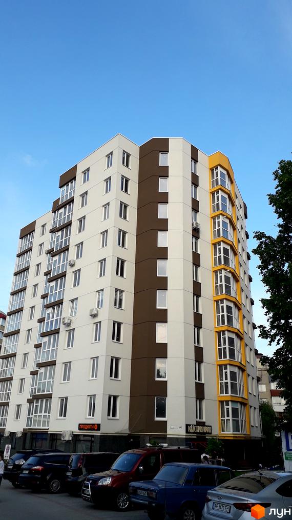 Багатоповерхова новобудова з білими та коричневими фасадами. На першому поверсі розташовані магазини, перед будинком припарковані автомобілі.