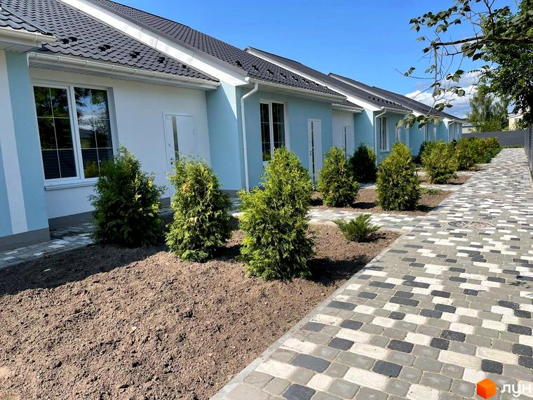 Ряд таунхаусів SMARTS зі світло-блакитними та білими фасадами, темними дахами. Перед будинками висаджені зелені кущі та прокладена бруківка.