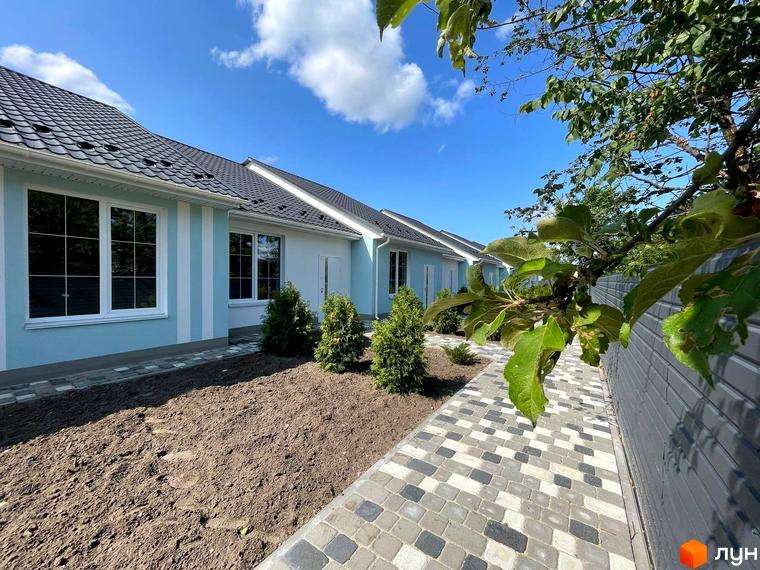 Ряд таунхаусів SMARTS зі світло-блакитними та білими фасадами, темними дахами та білими вікнами, поруч вимощена доріжка та підготовлені ділянки для озеленення.