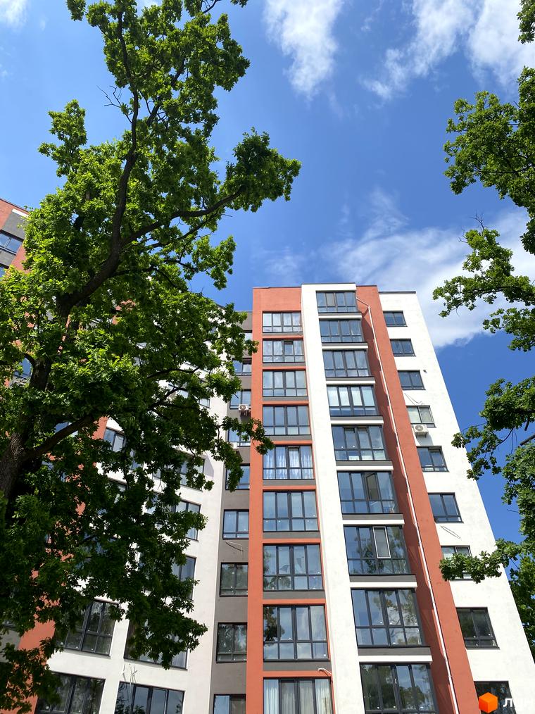 Житловий комплекс Green Life-3 має біло-помаранчевий фасад з численними вікнами. Навколо будівлі ростуть великі дерева з пишним зеленим листям.