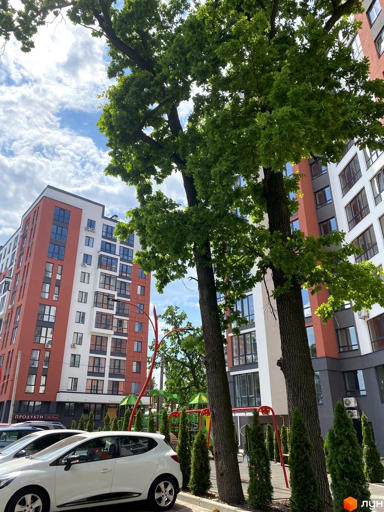 Новобудова ЖК Green Life-3 з червоно-білим фасадом та великими вікнами. На прибудинковій території розташований дитячий майданчик та зелені насадження.