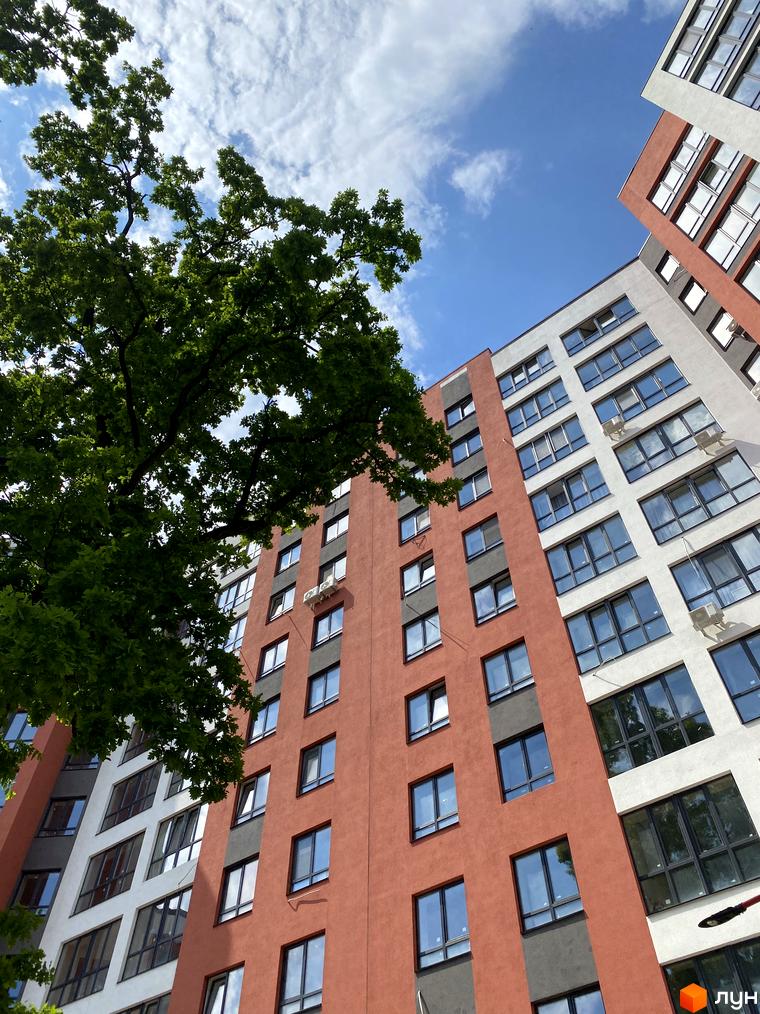 Фасад житлового комплексу Green Life-3 з червоними та білими елементами, вікнами та кондиціонерами. Навколо видно зелені дерева та блакитне небо з хмарами.