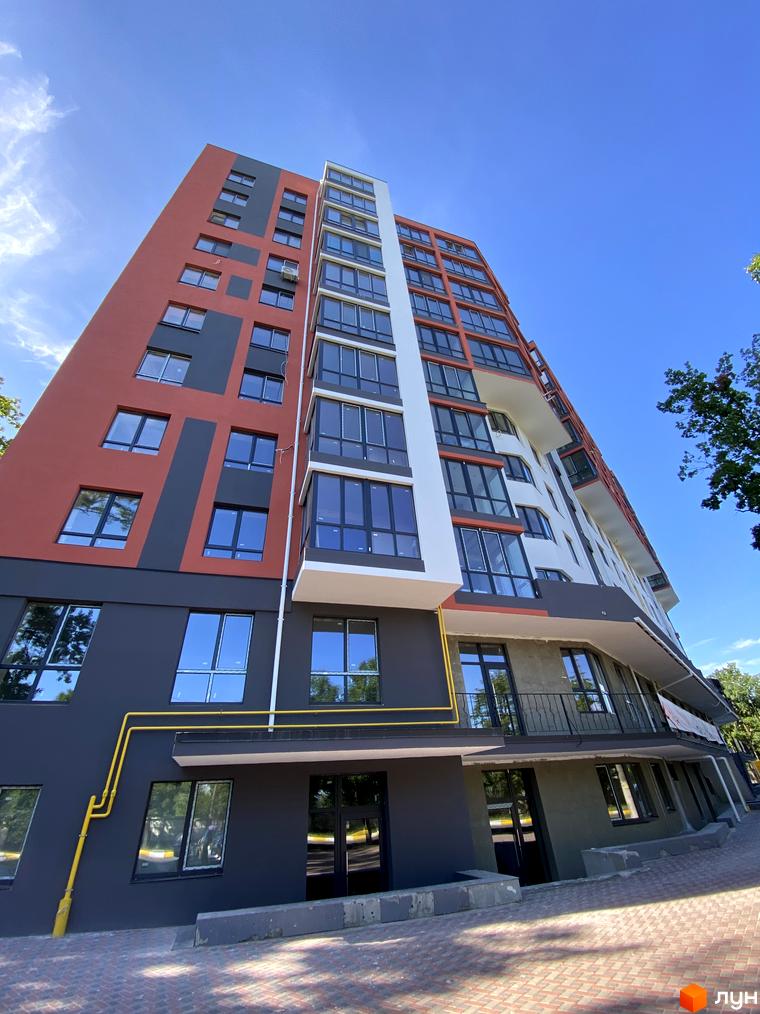 Висотна новобудова з фасадом червоного, сірого та білого кольорів. На нижньому поверсі будівлі розташовані великі вікна та двері, а також жовта газова труба. Поруч з будівлею викладена бруківка.