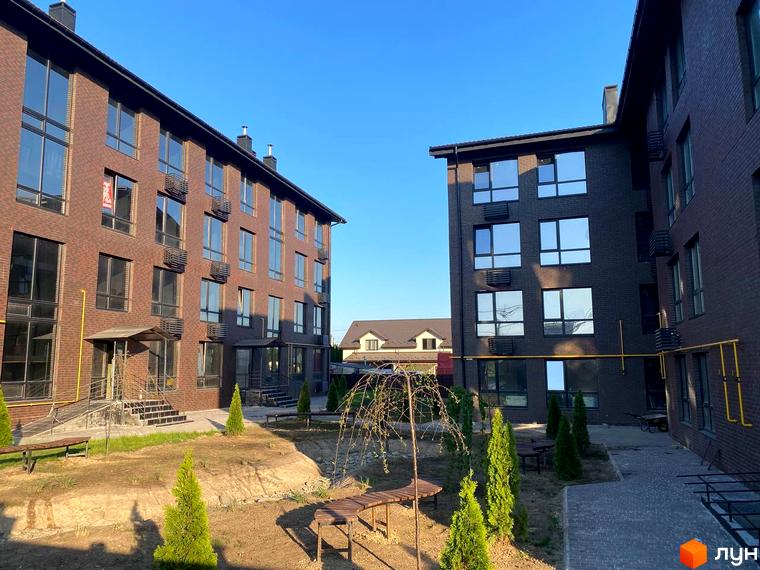 Зовнішній вигляд новобудови ЖК Моя Баварія з цегляним фасадом та великими вікнами. На прибудинковій території розташовані зелені насадження та лавки.