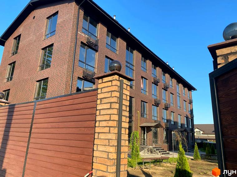 Зовнішній вигляд цегляної новобудови ЖК Моя Баварія з дерев'яним парканом та зеленими насадженнями на території.