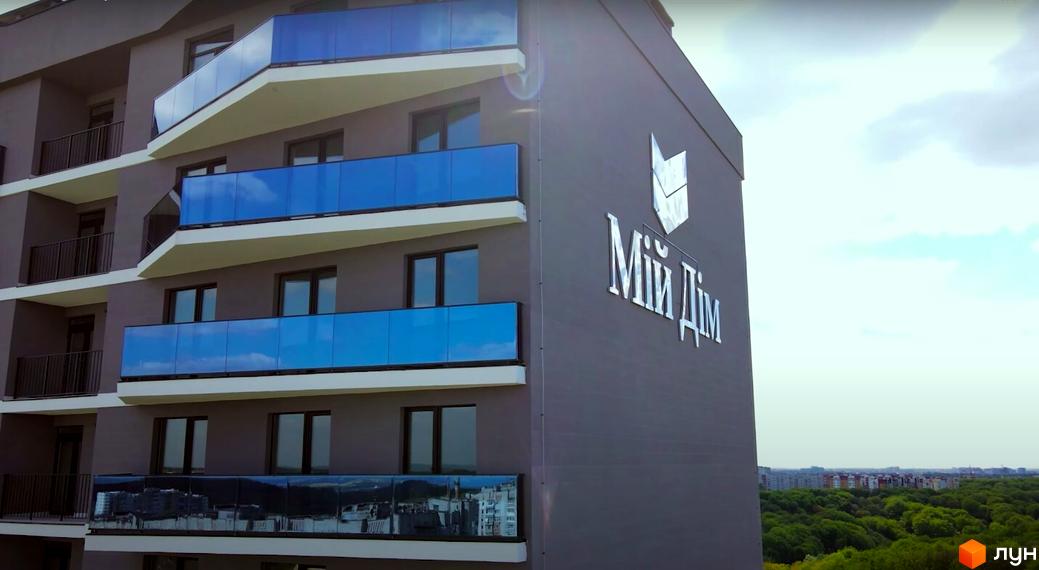 Фасад житлового комплексу Tiffany з блакитними скляними балконами та логотипом на стіні. Навколо зелені дерева та міський пейзаж.