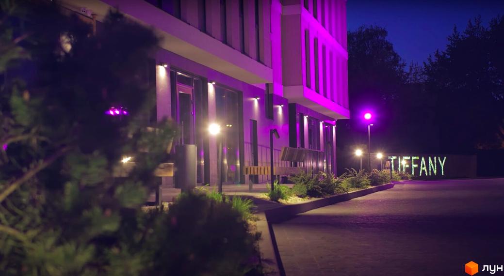 Житловий комплекс Tiffany з фасадом, підсвіченим фіолетовим світлом, та освітленою прибудинковою територією вночі.
