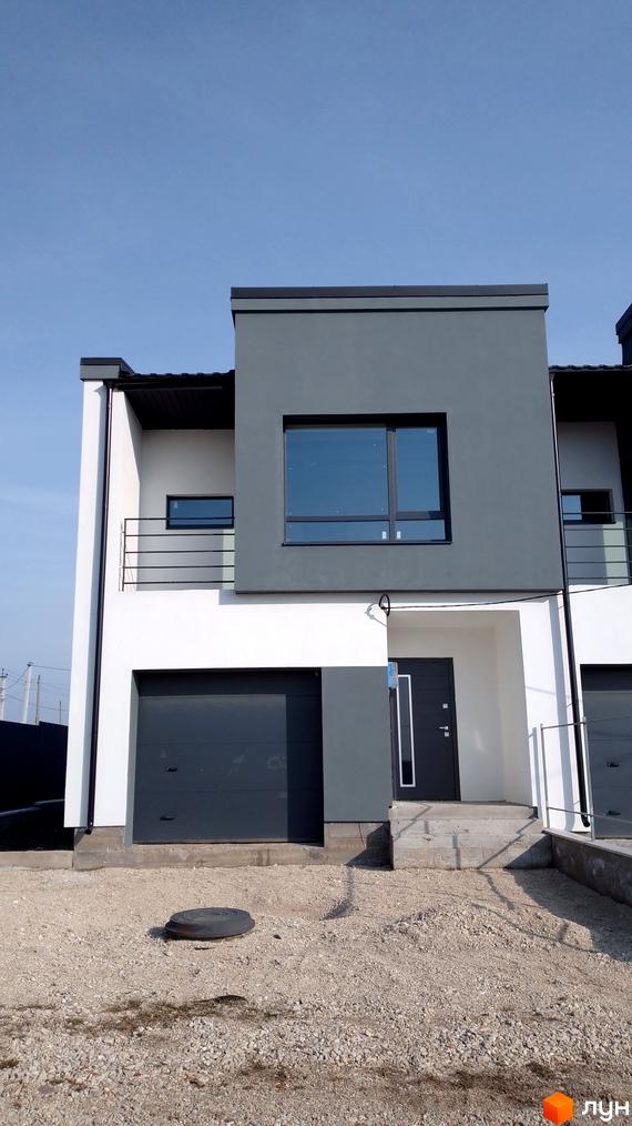 Фасад таунхауса Molodizhna Town з гаражем та балконом на тлі блакитного неба. Прибудинкова територія засипана гравієм.