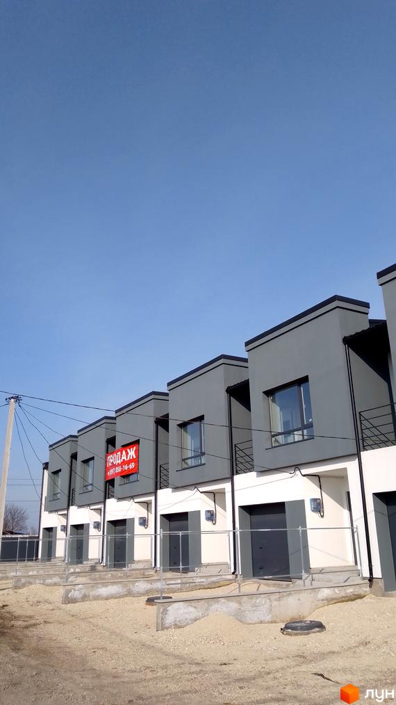Зовнішній вигляд таунхаусів Molodizhna Town з сірими фасадами та великими вікнами, на тлі блакитного неба. Перед будинками облаштована прибудинкова територія.